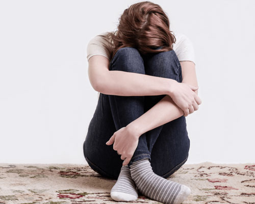 A teenage girl sitting on the floor with her back against a wall, holding her knees to her chest and looking anxious, illustrating panic attacks in teenagers.