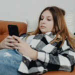 A teenage girl sitting on a couch with a worried expression, holding her mobile in her hands, illustrating generalized anxiety in teenagers.