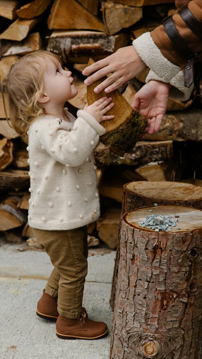 A toddler wearing a red shirt and blue jeans tries to lift a heavy log in a backyard, symbolizing the frequent accidents in toddlers due to their curiosity and developing motor skills.